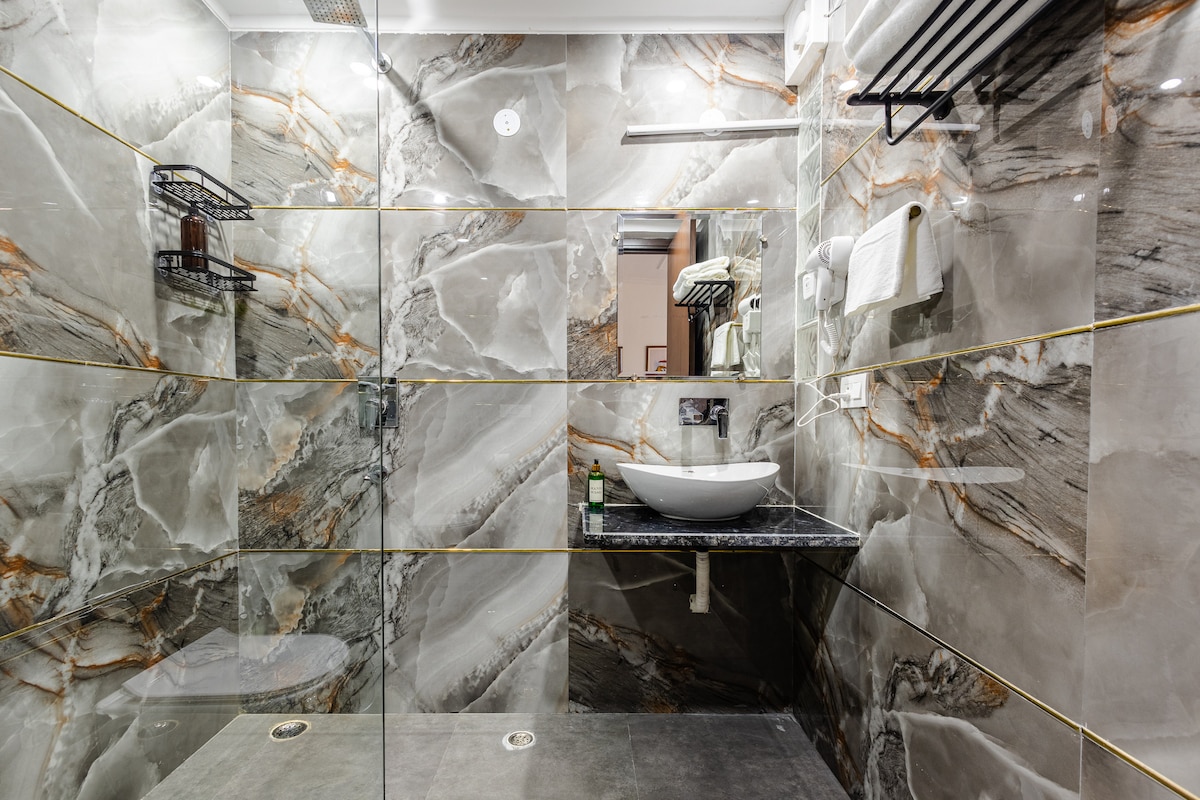 A modern bathroom features elegant marble walls with intricate veining in shades of grey and gold. A minimalist sink is mounted on a dark stone countertop, and a large mirror reflects the stylish design. Subtle lighting enhances the luxurious ambiance.