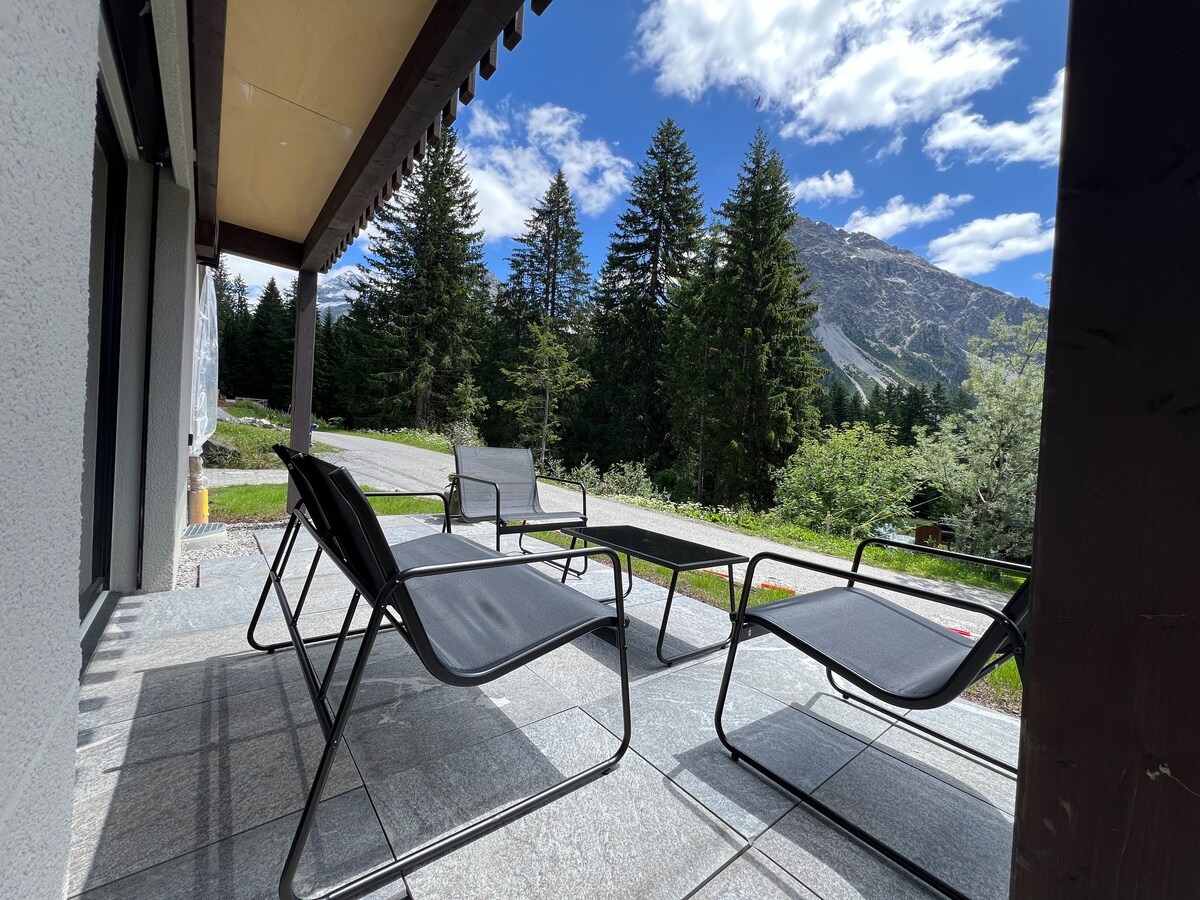 A shaded seating area is present on a stone terrace, featuring two lounge chairs and a coffee table. The view includes lush greenery and tall trees, with mountains rising in the background under a partly cloudy sky.