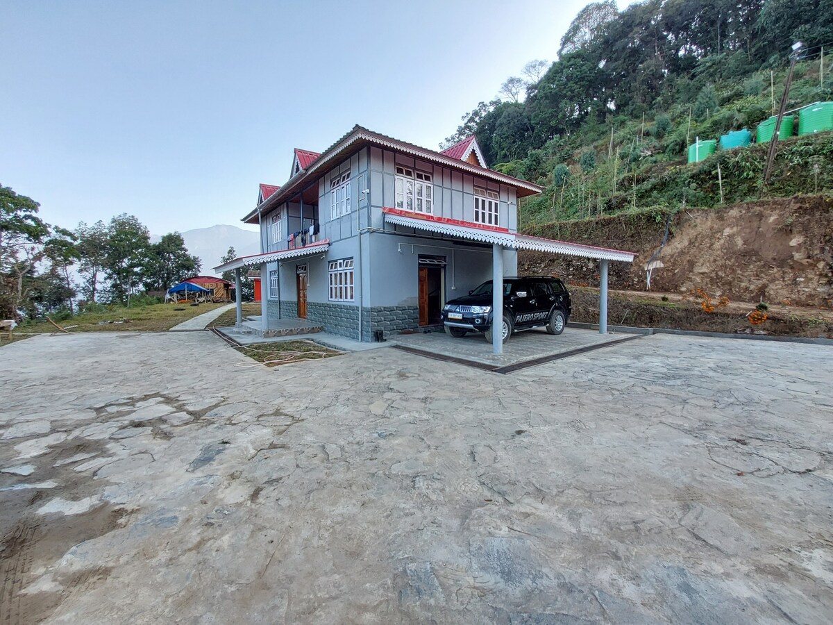 A two-story farmhouse is positioned on a spacious stone driveway, surrounded by lush greenery. The exterior features a light gray facade with red accents on the roof. A parked car is visible beneath a covered porch, indicating easy access for guests.