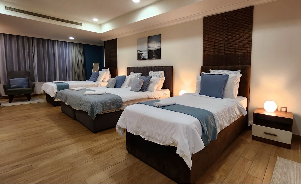 A spacious master bedroom with two king size beds is presented, alongside a cozy armchair. Soft furnishings in muted tones are complemented by natural light filtering through sheer curtains. Warm wood flooring enhances the overall comfort of the inviting space.