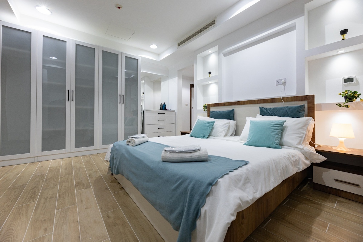 A spacious bedroom features a comfortable bed with light blue accents and neatly arranged white linens. Built-in wardrobes with glass doors provide ample storage. Soft lighting creates a calming ambiance, while wooden floors add warmth to the modern design.