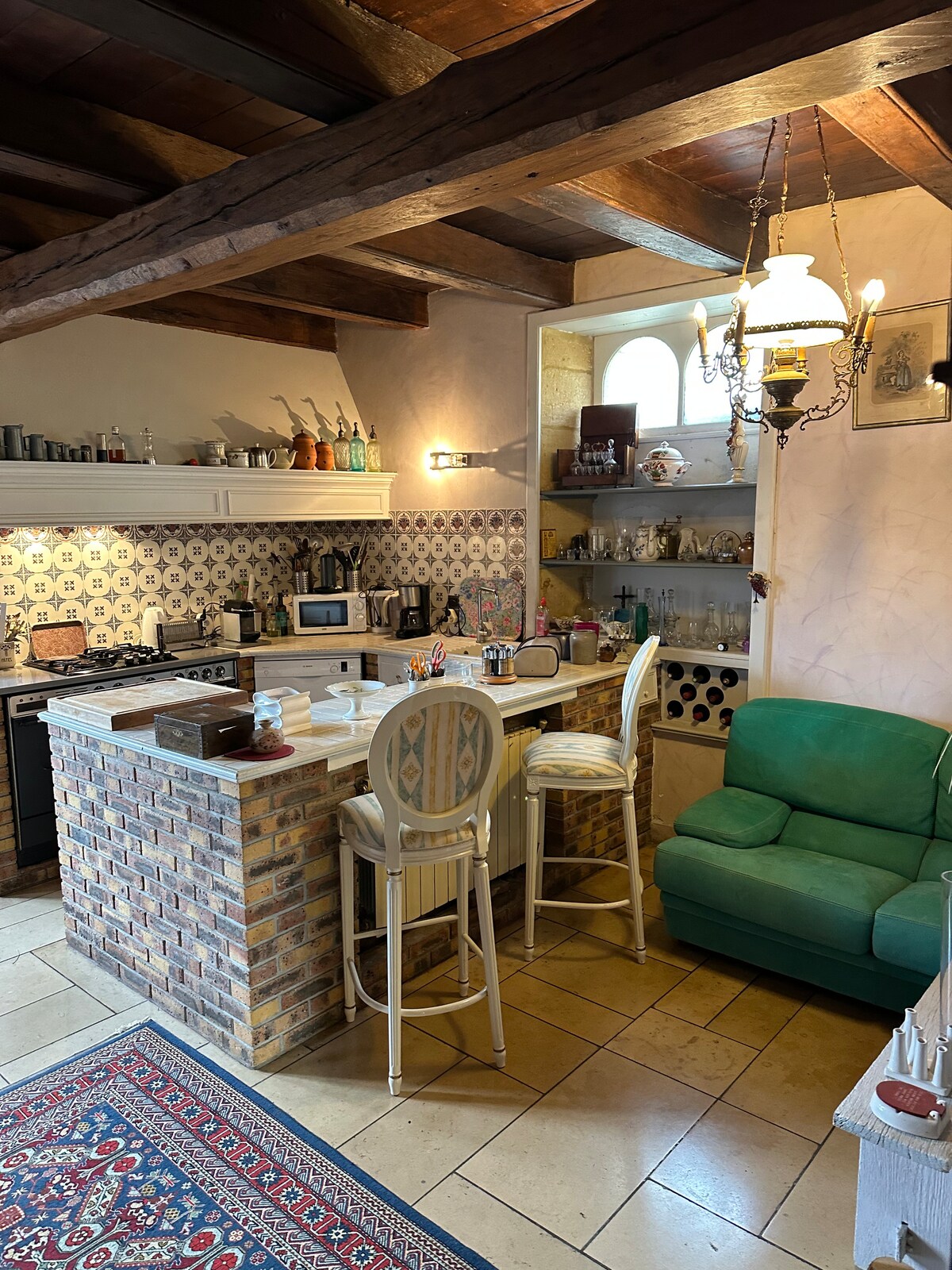 A cozy kitchen space features exposed wooden beams and classic tile work. A small breakfast bar with white stools is visible, alongside modern appliances and glass shelves displaying dishware. A green armchair adds a pop of color, and an area rug complements the tiled floor.