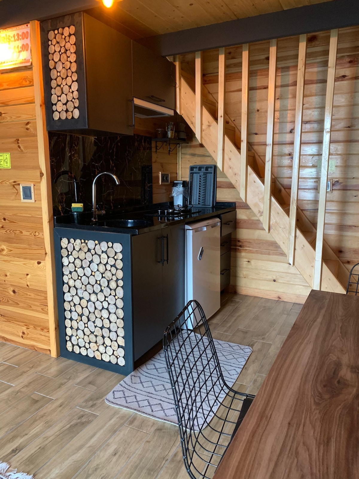 An open kitchen area showcases modern appliances, including a gas stove and a refrigerator. Natural wood accents adorn the walls, with a wooden dining table set nearby. A textured rug lies on the floor, complementing the contemporary design of the wire dining chairs.