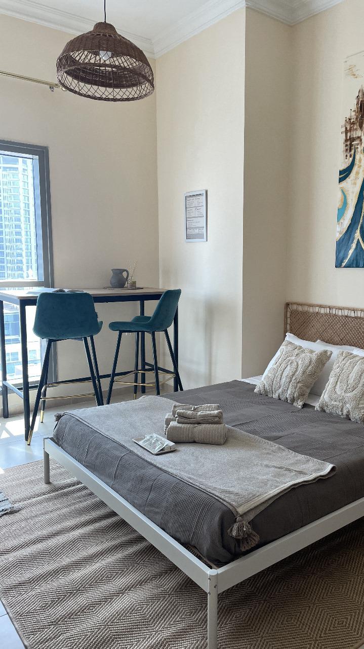 A cozy sleeping area features a spacious bed dressed in neutral tones. Light gray linens complement decorative pillows. A small dining table with two blue chairs is visible near a window, creating a bright and airy ambiance within the room.