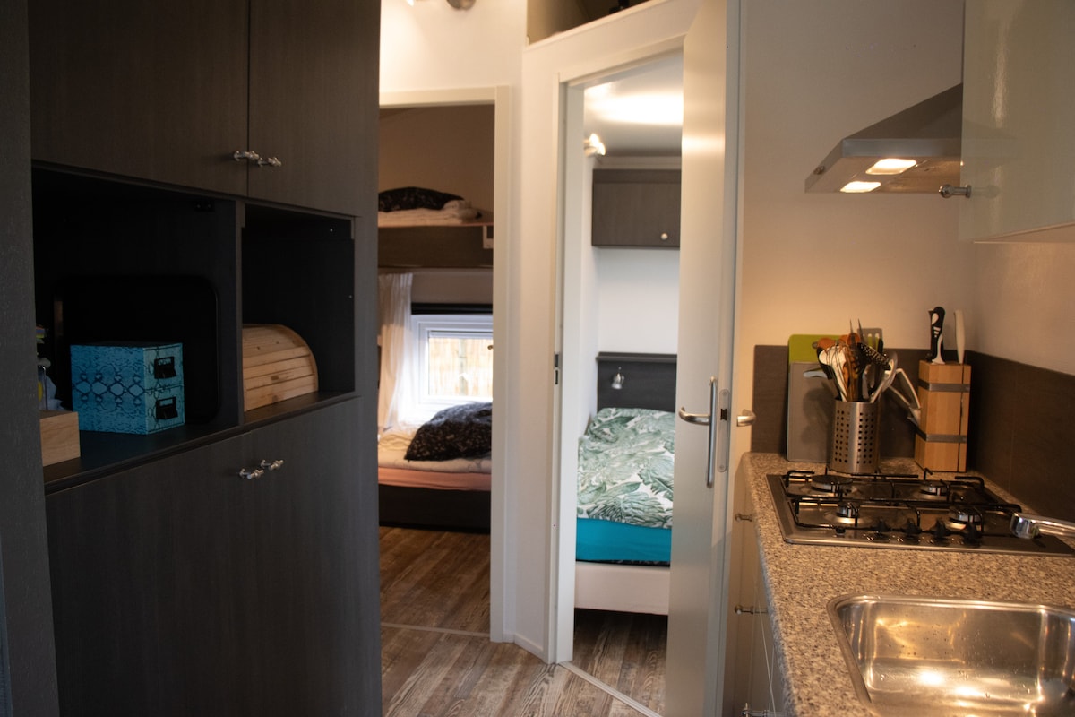 The image features a view of the kitchen area leading to bedrooms, highlighted by a gas stove and a sleek countertop. Shelving units are visible, providing storage space. The entrance to a bedroom is seen, with a bunk bed in the background and a bed with bedding in another bedroom.
