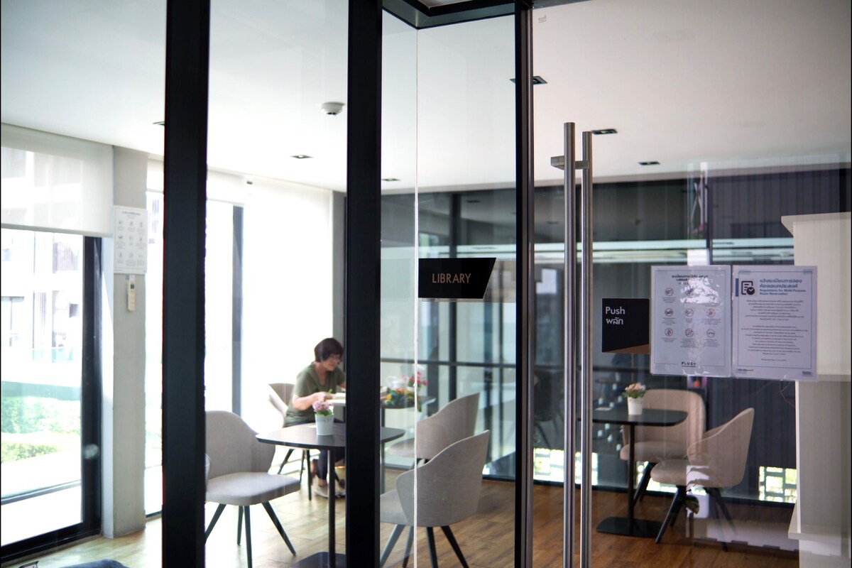 The image depicts a modern co-working space with glass walls. Inside, white chairs surround dark tables, and natural light illuminates the area. A person is seated at one of the tables, engaged in work, while a clear sign indicates the entrance to the library.