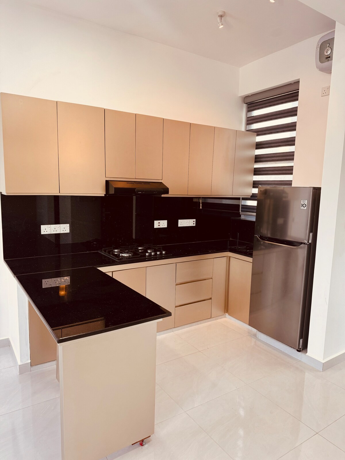 A modern kitchen is equipped with sleek beige cabinets and a dark granite countertop. A gas stove and built-in oven are integrated, while a silver refrigerator stands beside. Natural light filters through striped window coverings, enhancing the space's clean, contemporary aesthetic.