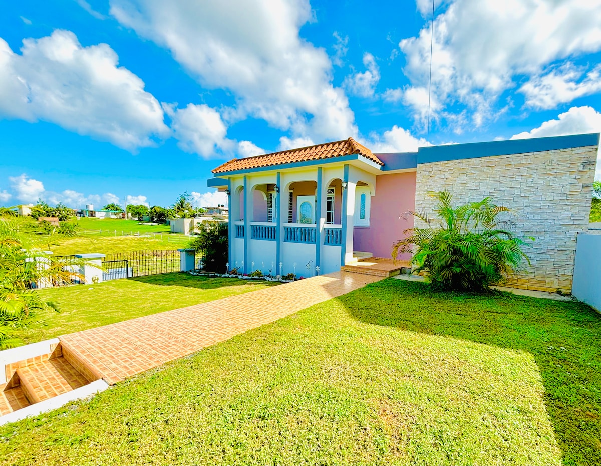 A charming exterior showcases a single-story home featuring a covered porch with arched openings. Lush green grass and landscaped gardens surround the property, complemented by a blue sky dotted with fluffy clouds.