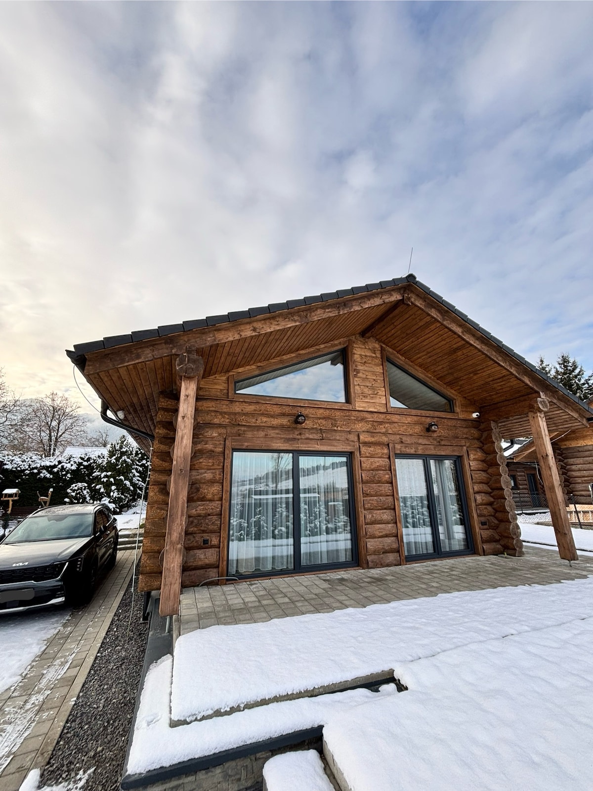 The exterior of a wooden chalet is showcased, featuring large glass doors that open to a snow-covered pathway. The roofline is angular, complemented by natural wood pillars. The surrounding area is serene, with light snow blanketing the ground and a modern car parked nearby.