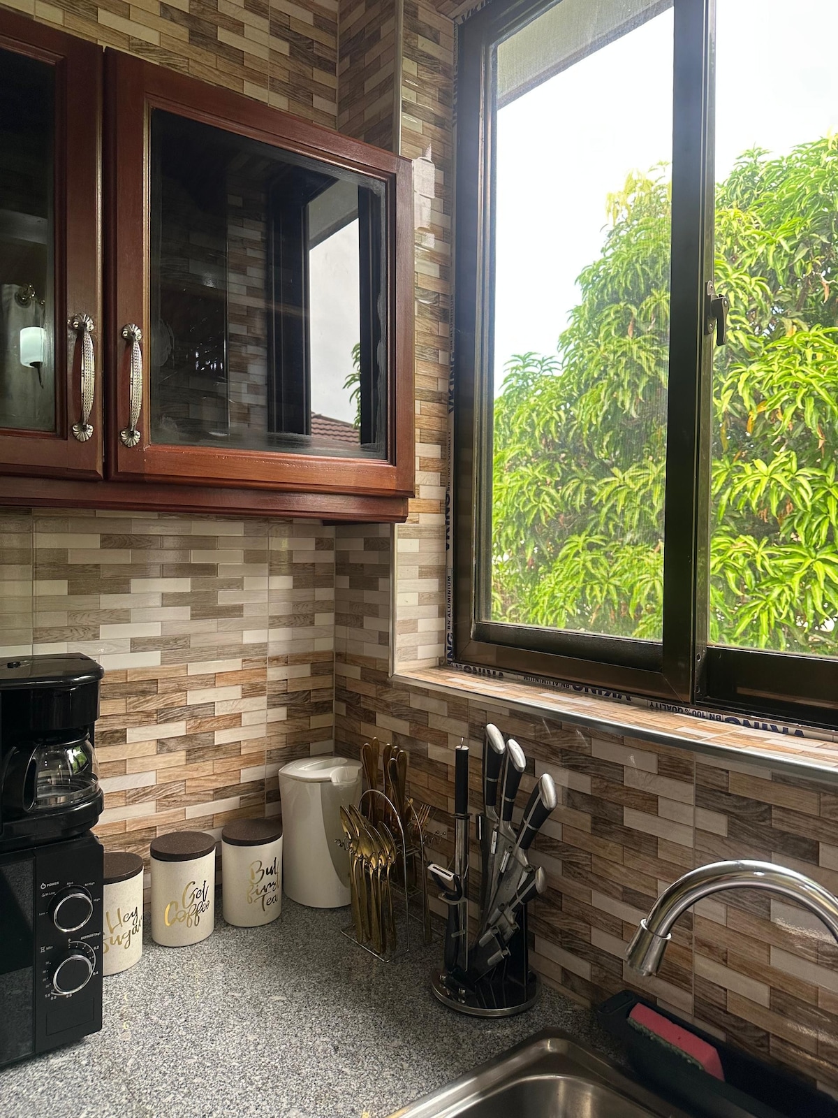 A kitchen corner features a countertop with a coffee maker, ceramic storage containers, and a collection of utensils in a holder. Natural light enters through a window, highlighting the mosaic tiled wall and cabinetry with glass-front doors.