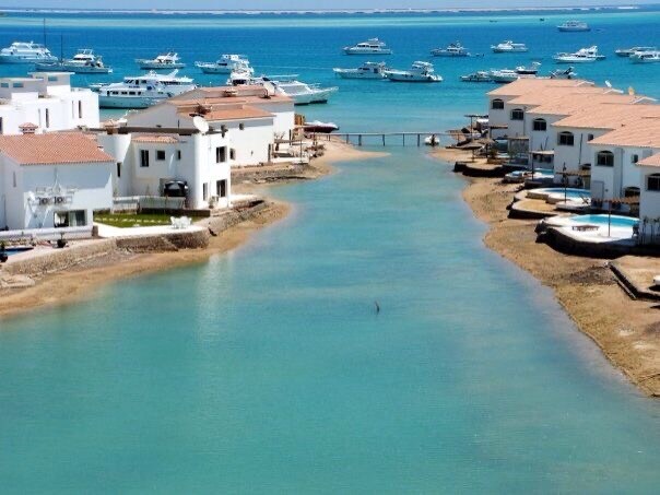 A serene waterway flows between white buildings with terracotta rooftops, leading to a picturesque lagoon. Moored boats are visible in the calm blue waters, creating a relaxing coastal environment. The scene conveys a sense of tranquility and connection to the sea.