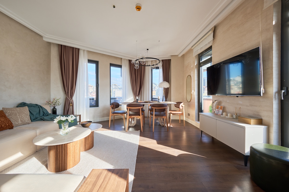 A spacious living area is highlighted by large windows, allowing natural light to fill the room. A comfortable sofa and wooden coffee tables are present, complemented by a dining table surrounded by four chairs. Decorative elements and a television add to the modern decor.