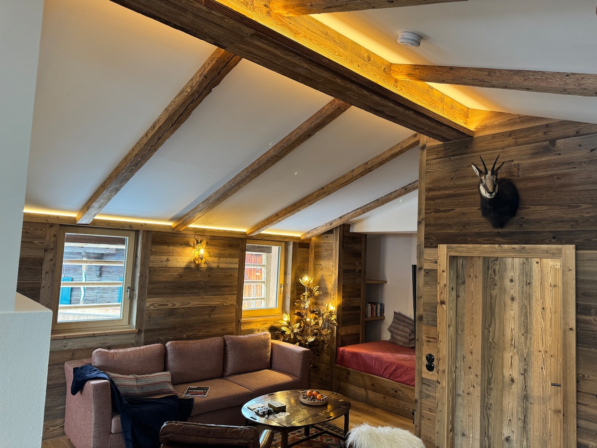 A cozy living space is characterized by wooden beams and warm lighting. A soft sofa is positioned near a coffee table, while a sleeping area features a bedside with neatly arranged linens. Two windows allow natural light to fill the room, enhancing the rustic decor.