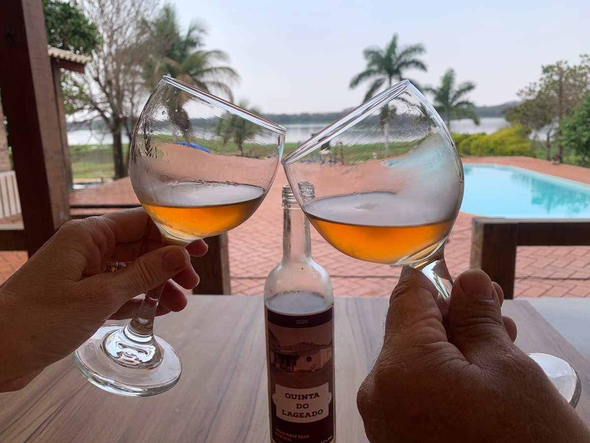 Two glasses are raised in a toast, showcasing a warm beverage. In the background, a serene swimming pool is visible, complemented by lush greenery and palm trees. A bottle of wine labeled 'Quinta do Lagedo 2020' rests on the table.