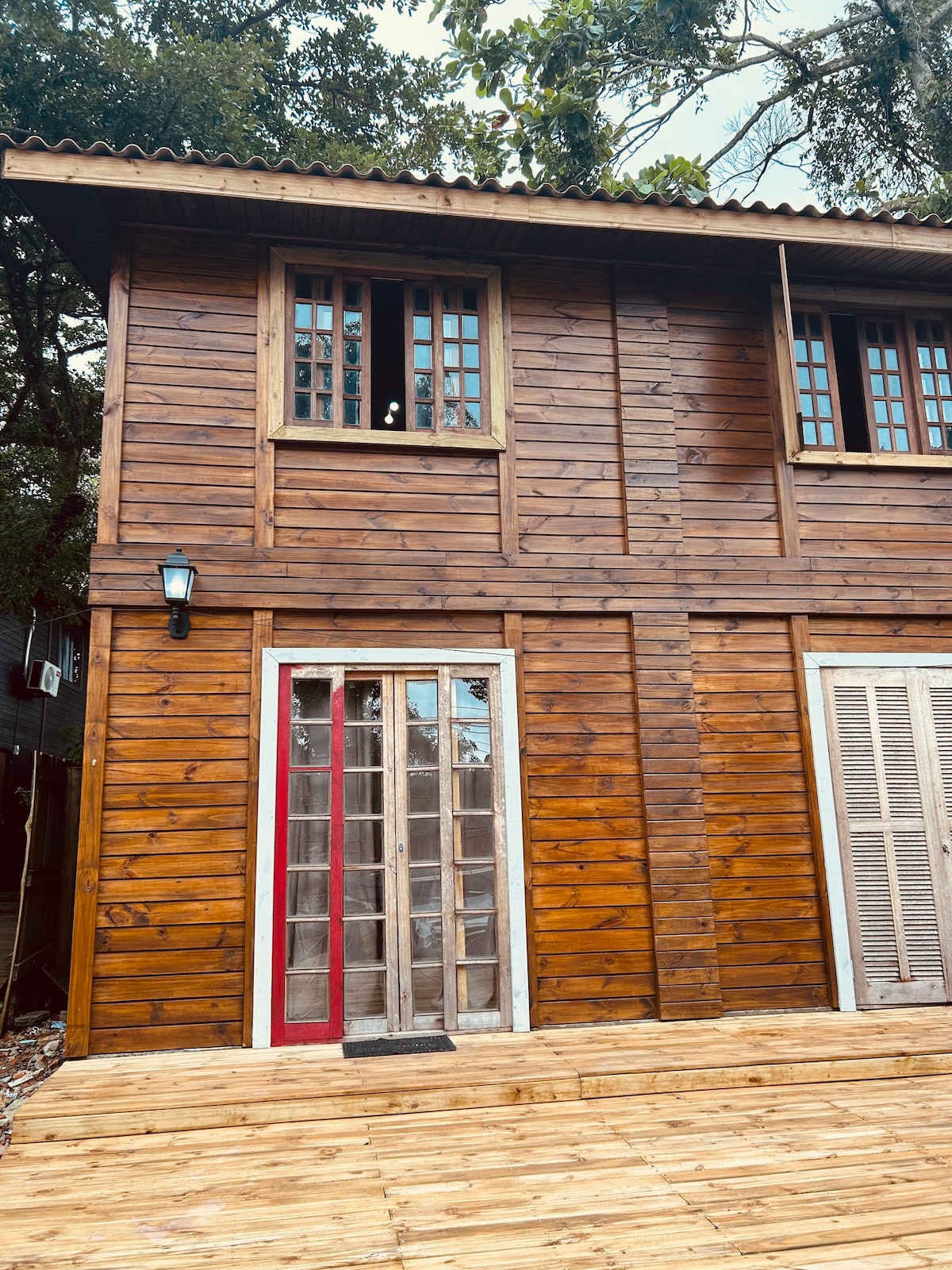 The exterior view of a two-story wooden house reveals a warm, rustic design. Large glass doors provide access to the ground level, framed by wooden siding. Multiple windows with wooden frames allow natural light to illuminate the interior while a porch extends in front.