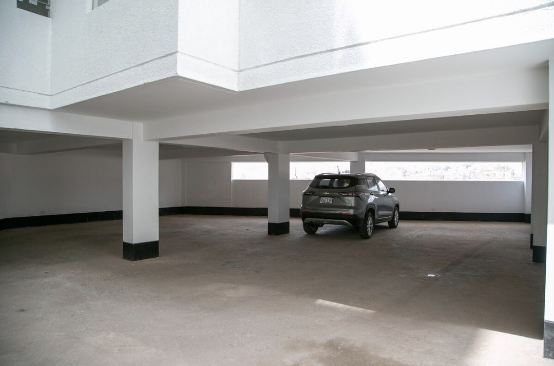 A spacious parking area is shown with clean, white walls and a smooth concrete floor. Natural light streams in through openings on the far wall, highlighting a parked vehicle situated in a designated space.