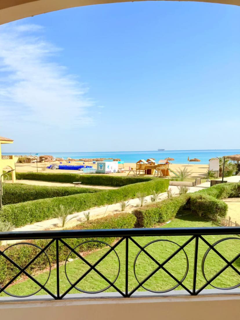 A view from a balcony showcases a sandy beach lined with lounge chairs and recreational equipment. Lush green landscaping frames the scene, while the deep blue sea stretches toward the horizon under a clear sky.