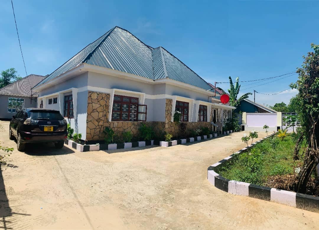A spacious single-story building is shown with a distinctive stone exterior and a metal roof. A well-maintained driveway leads up to the entrance, flanked by greenery. Ample parking space is available in front of the house, adding to its welcoming appearance.