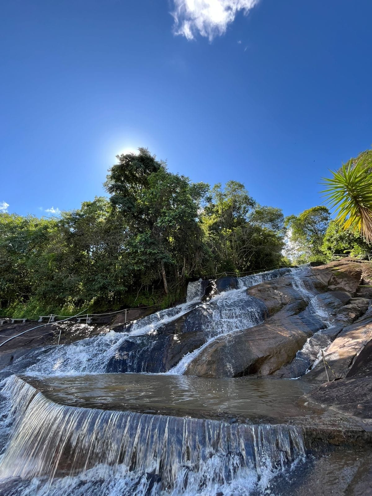 A cascading waterfall tumbles down smooth rocks, creating small pools of clear water at the base. Lush greenery surrounds the area, with tall trees and plants visible, set against a bright blue sky with a few fluffy clouds.