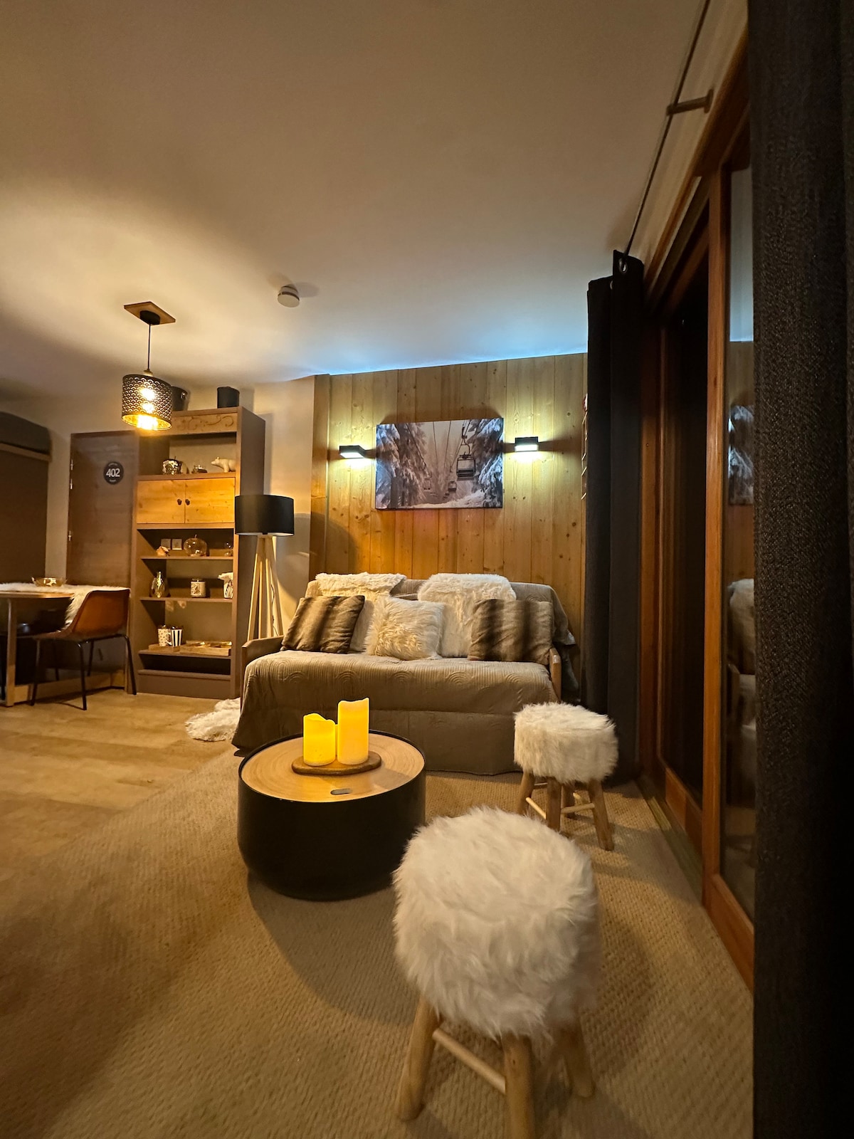 A cozy sitting area features a plush sofa adorned with soft cushions, set against a warm wooden wall. A circular table with decorative candles is accompanied by two fur-covered stools. Dim lighting creates a relaxed atmosphere, and a display shelf with various items adds to the decor.
