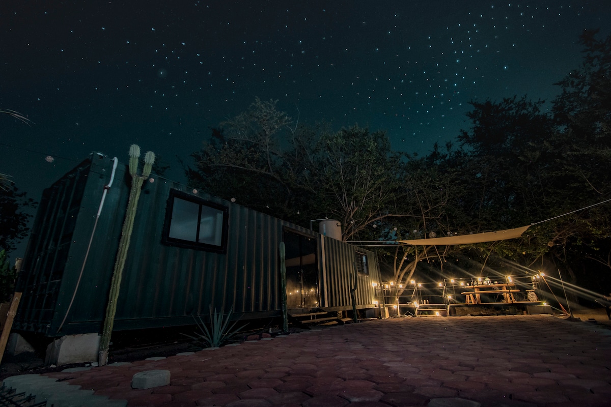 A rustic container accommodation is nestled under a starry sky, surrounded by trees. Soft lighting from string lights enhances the outdoor dining area, which includes a picnic table. A shaded area is visible, supported by a canopy, creating a cozy retreat in nature.