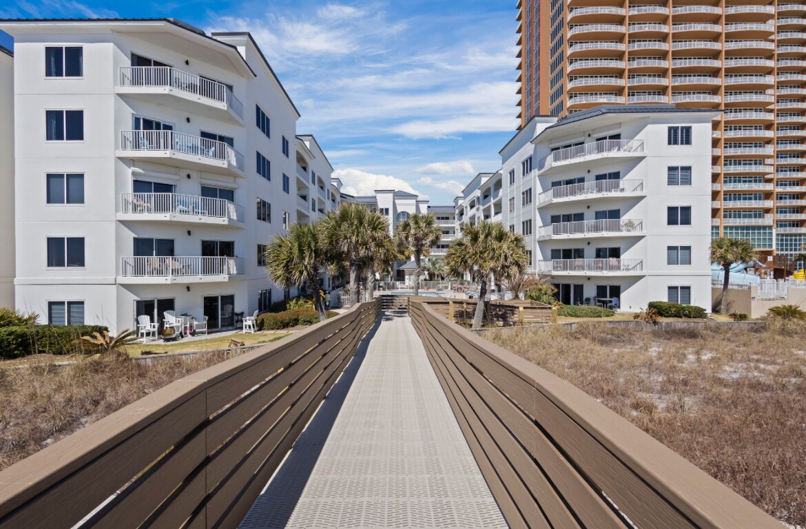 A broad boardwalk leads through landscaped grounds towards beach access, framed by modern white buildings on either side. Lush palm trees provide natural scenery, while the backdrop features a tall structure, indicative of the coastal environment.