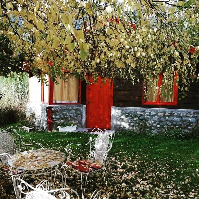A quaint cabin is framed by overhanging branches, with a scattering of yellowed leaves. A circular table and four metal chairs sit on the grassy area in front, surrounded by more fallen leaves. The cabin features red accents and is partially shrouded by a stone wall.