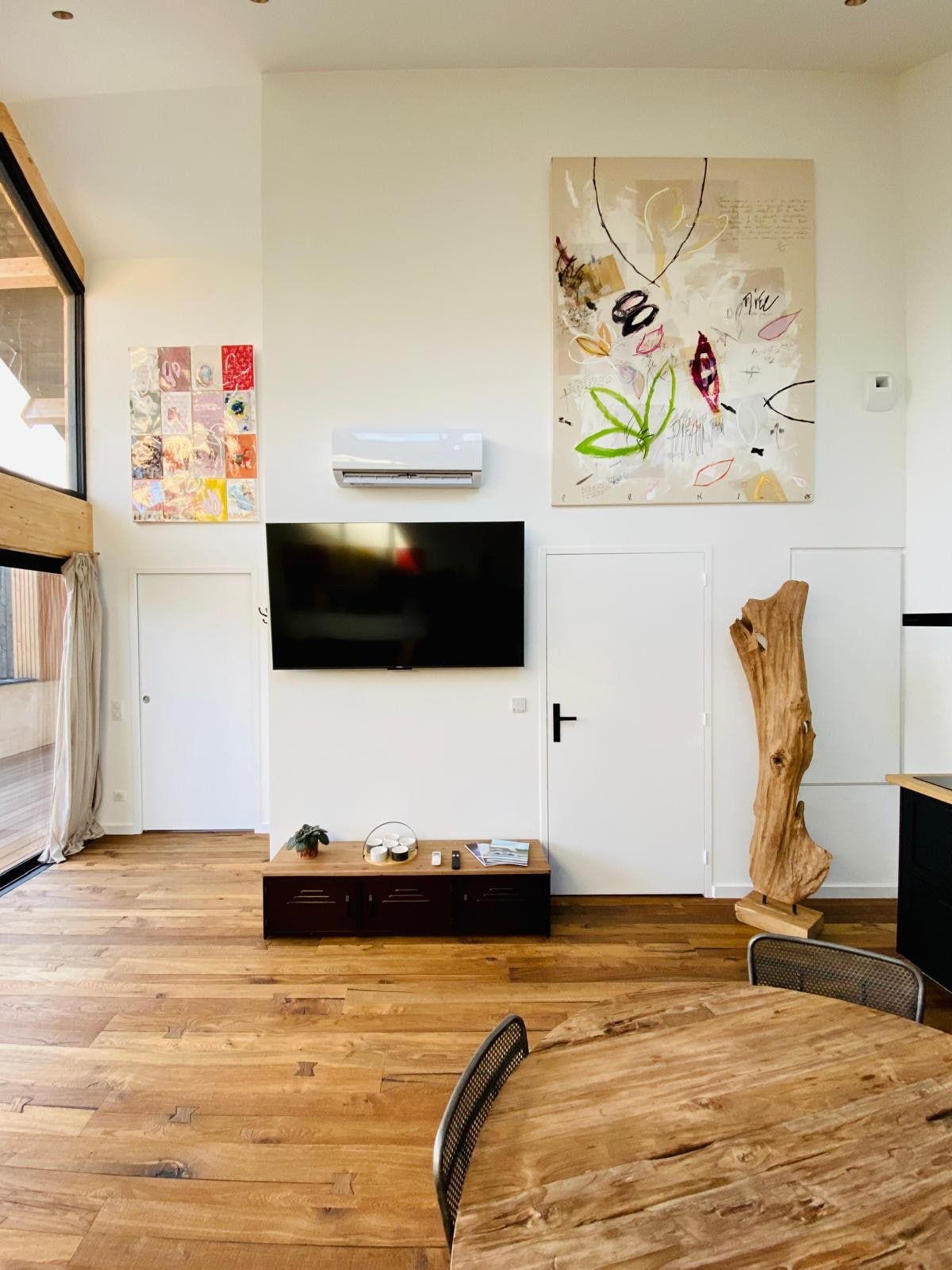 The living area features high ceilings with large windows that allow natural light to illuminate the space. Contemporary artwork decorates the walls, and a wooden table stands nearby. A flat-screen TV is mounted on the wall, complemented by a natural wood accent piece.