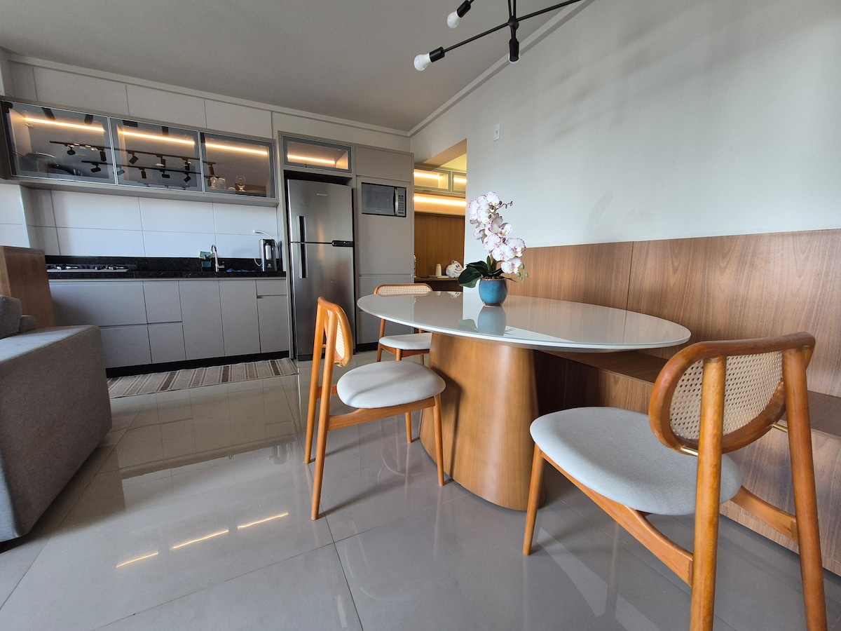 A modern dining area features a round table with wooden legs and three chairs. The space is highlighted by a light-colored cabinet and a kitchenette in the background. A potted plant adds a touch of greenery, complementing the sleek design.