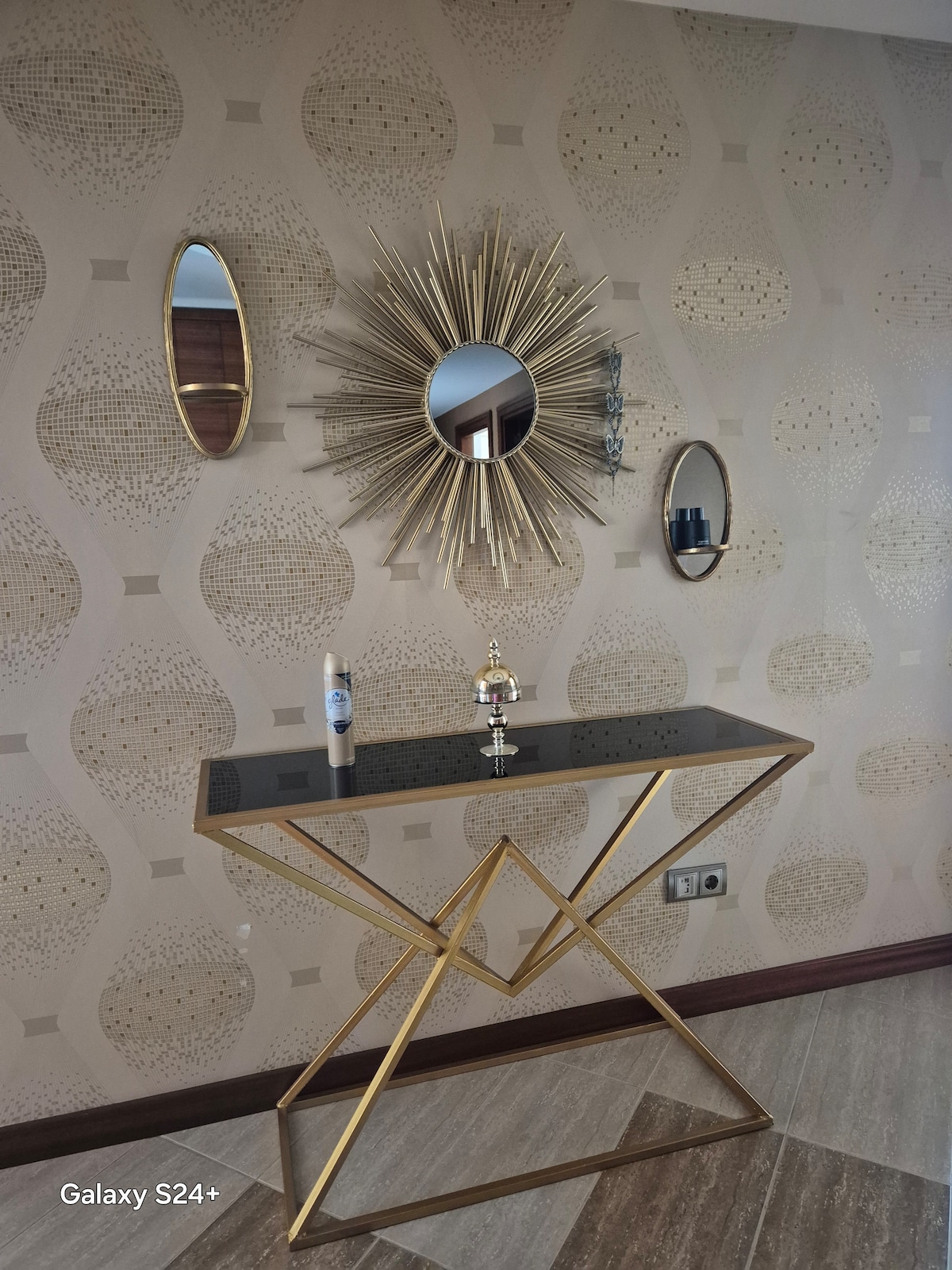 A stylish console table features a black surface and an elegant gold base, positioned against a patterned wall. Above it are three mirrors, including a sunburst design, reflecting light and enhancing the space. Two decorative items add to the overall aesthetic.