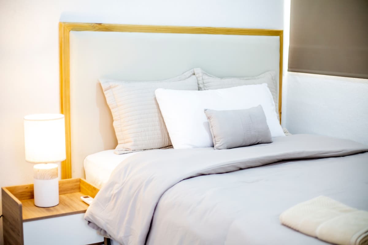 A cozy sleeping area features a light-colored bed with an upholstered headboard. Soft pillows, including a decorative one, are arranged on the bed. A bedside table with a lamp illuminates the space, which is complemented by a light-colored blanket.