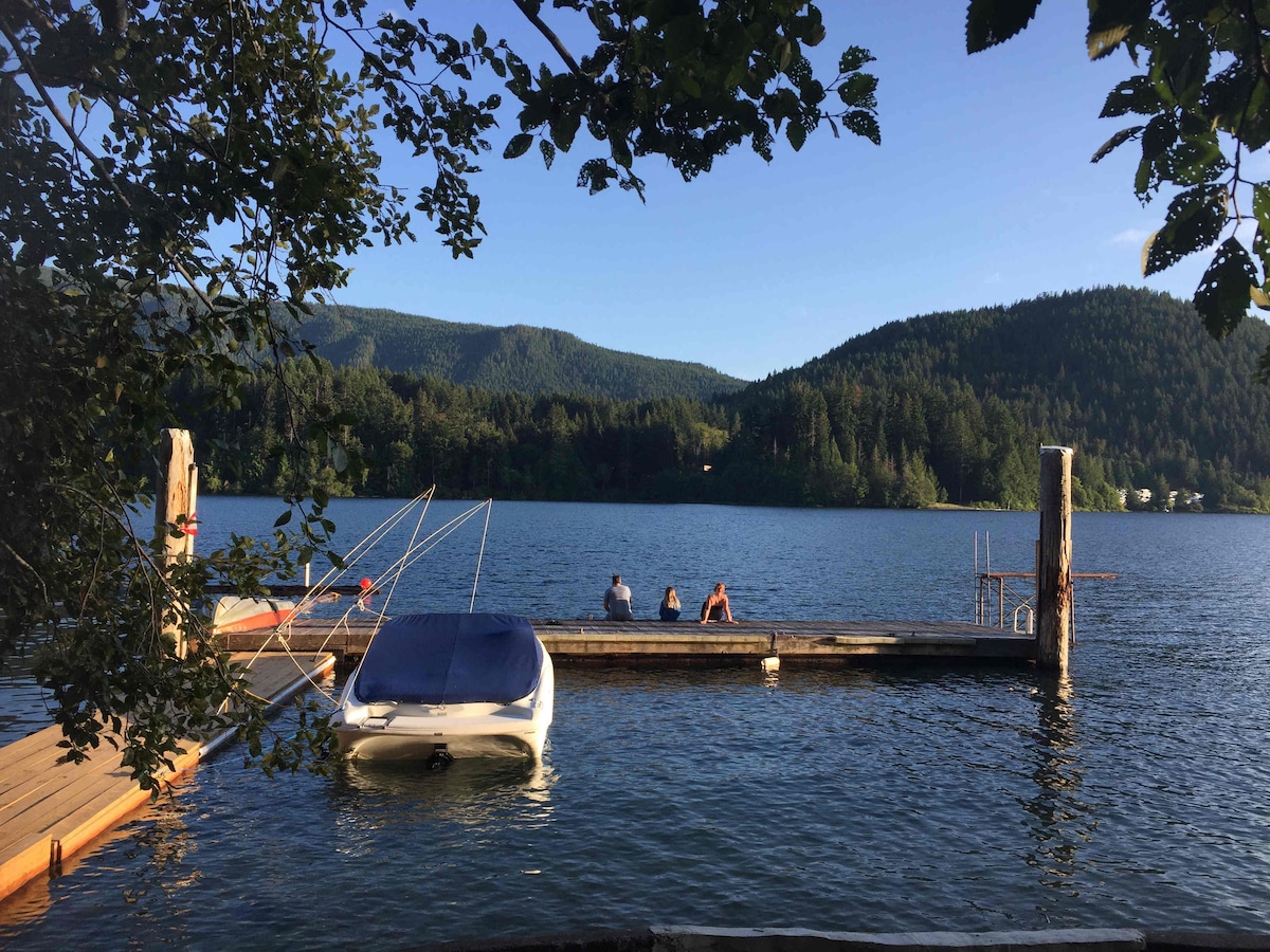 A wooden dock extends over calm waters, with three individuals seated at the edge, enjoying the serene lake view. A small boat is moored beside the dock, framed by lush, green trees and distant mountains under a clear blue sky.