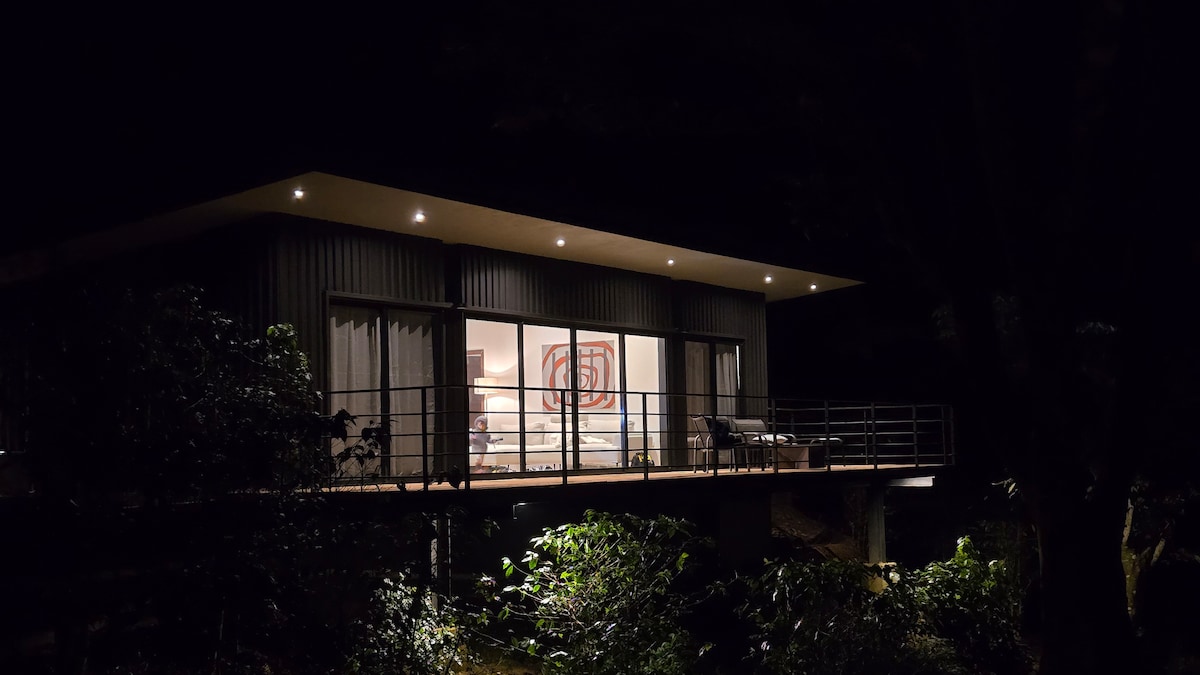 A modern cabin sits elevated among trees, featuring large glass windows that reflect warm interior lighting. A balcony extends outwards, complemented by a simple railing and a couple of outdoor chairs. Lush greenery surrounds the structure, contributing to the tranquil ambiance.