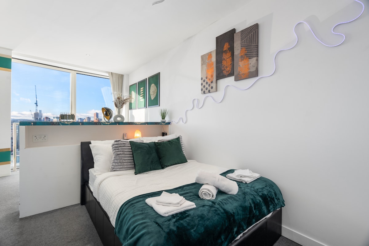 The sleeping area features a spacious bed with soft bedding and complementary decorative pillows. Artwork is displayed above, while a strip of LED lights provides ambient illumination. Natural light enters through a large window, accentuating the sleek, modern design of the room.