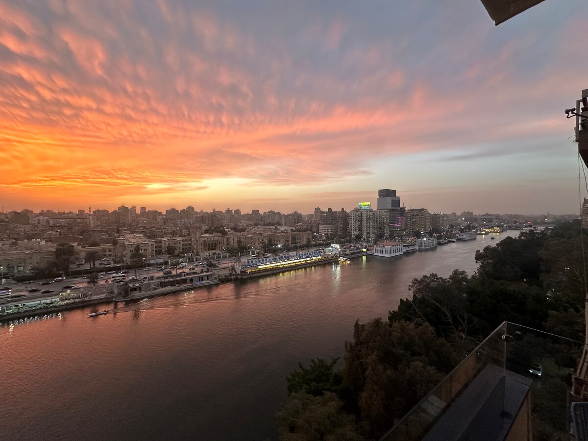 A panoramic view captures the Nile River at sunset, showcasing vibrant clouds in shades of orange and pink. City buildings line the riverbank, and boats are visible on the water, reflecting the colors of the sky in a serene urban landscape.