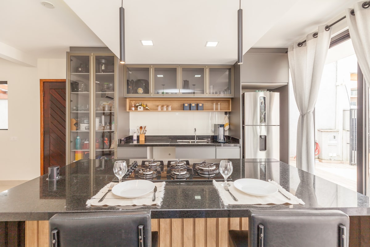 A spacious kitchen features a modern design with a large black granite island. Cooking appliances and utensils are neatly arranged on shelves behind the island, complemented by a sleek stainless steel refrigerator. Natural light fills the area through large windows, enhancing the welcoming atmosphere.