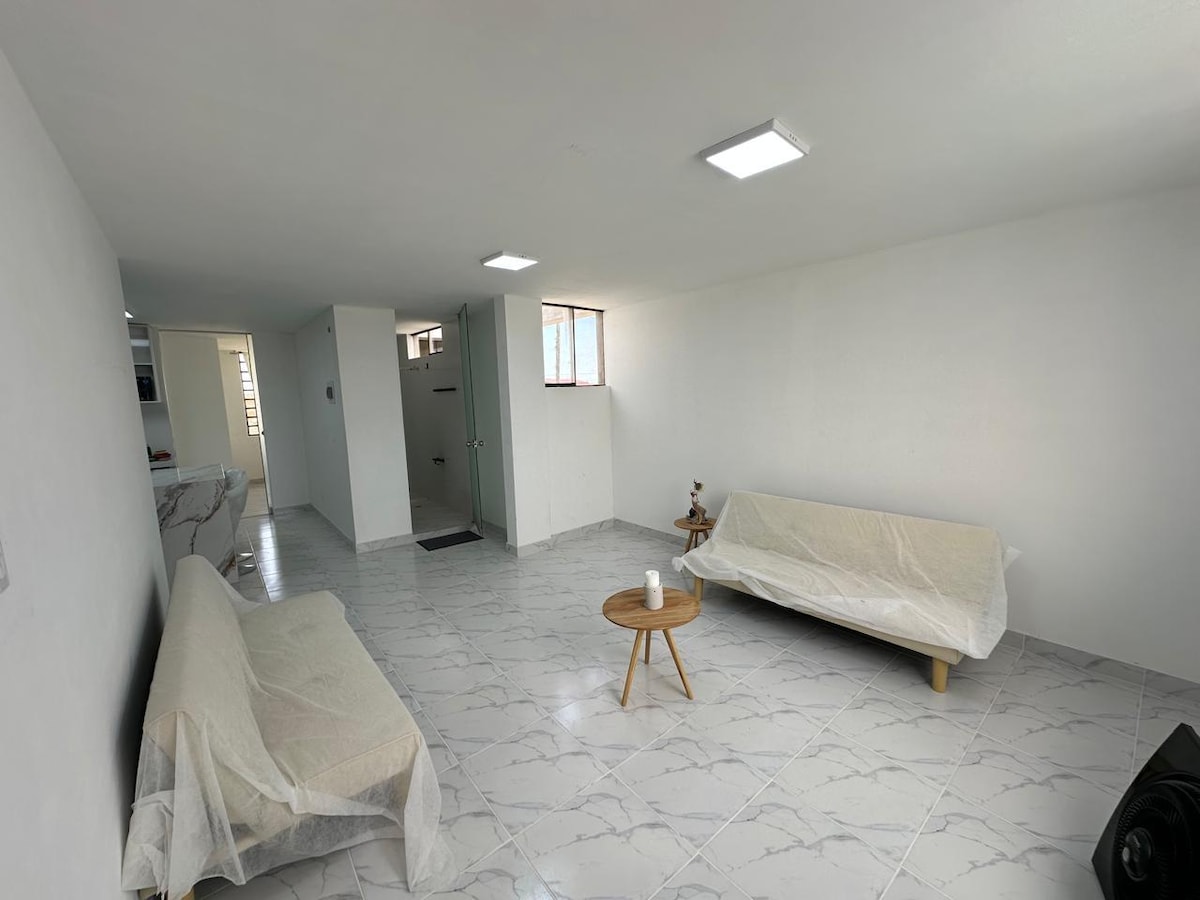 A spacious living area is presented with light-colored walls and glossy tile flooring. Two covered sofas are positioned around a round wooden coffee table. Natural light filters in through a window, illuminating the open space that connects to the layout of the departamento.