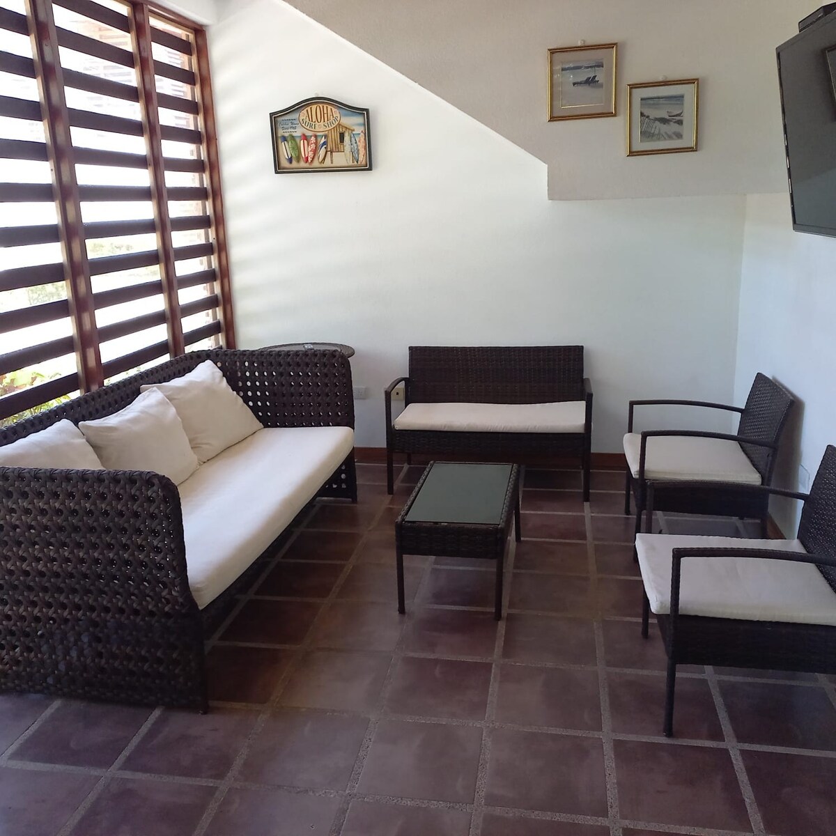 A comfortable lounge area features a woven seating arrangement with a spacious sofa and four chairs. A glass-top table is centrally placed. Natural light filters through wooden slats, creating a relaxed ambiance. Three framed artworks adorn the wall, adding subtle décor.