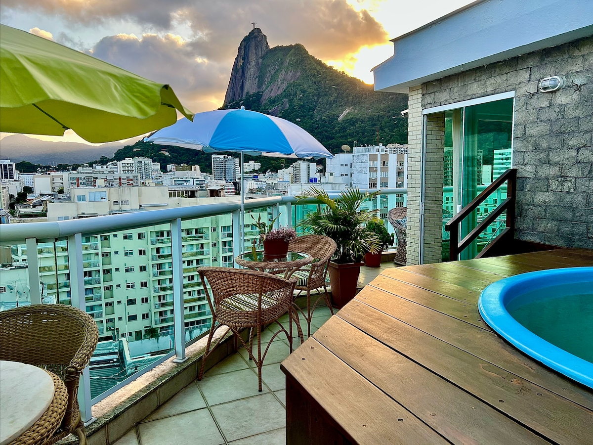 The terrace features a wooden hot tub surrounded by colorful umbrellas and stylish seating. Views of the mountains and cityscape are visible in the background, framed by a sunset sky. Potted plants add a natural touch to the outdoor space.