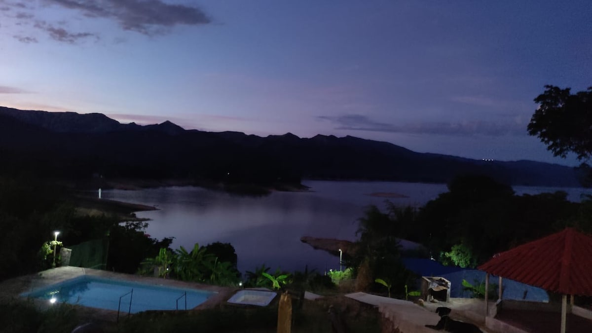 A serene view of a calm lake is presented at dusk, with the silhouette of mountains in the background. Soft lighting illuminates the surrounding area, and lush greenery can be seen along the water's edge, enhancing the tranquil ambiance.
