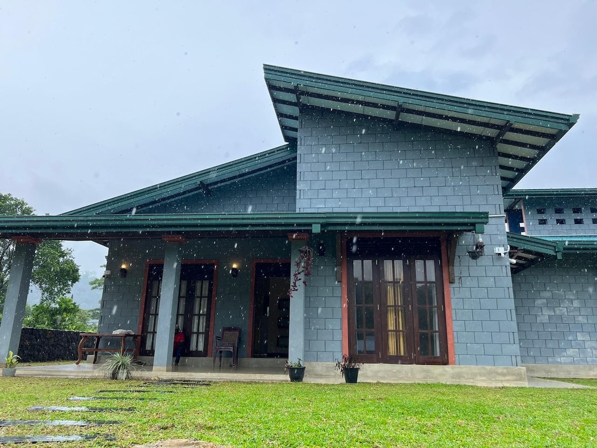 An inviting home with a grey stone exterior and sloped green roof is shown. Large glass doors lead to the entrance, framed by decorative plants. Lush green grass surrounds the property, creating a refreshing atmosphere even in light rain.