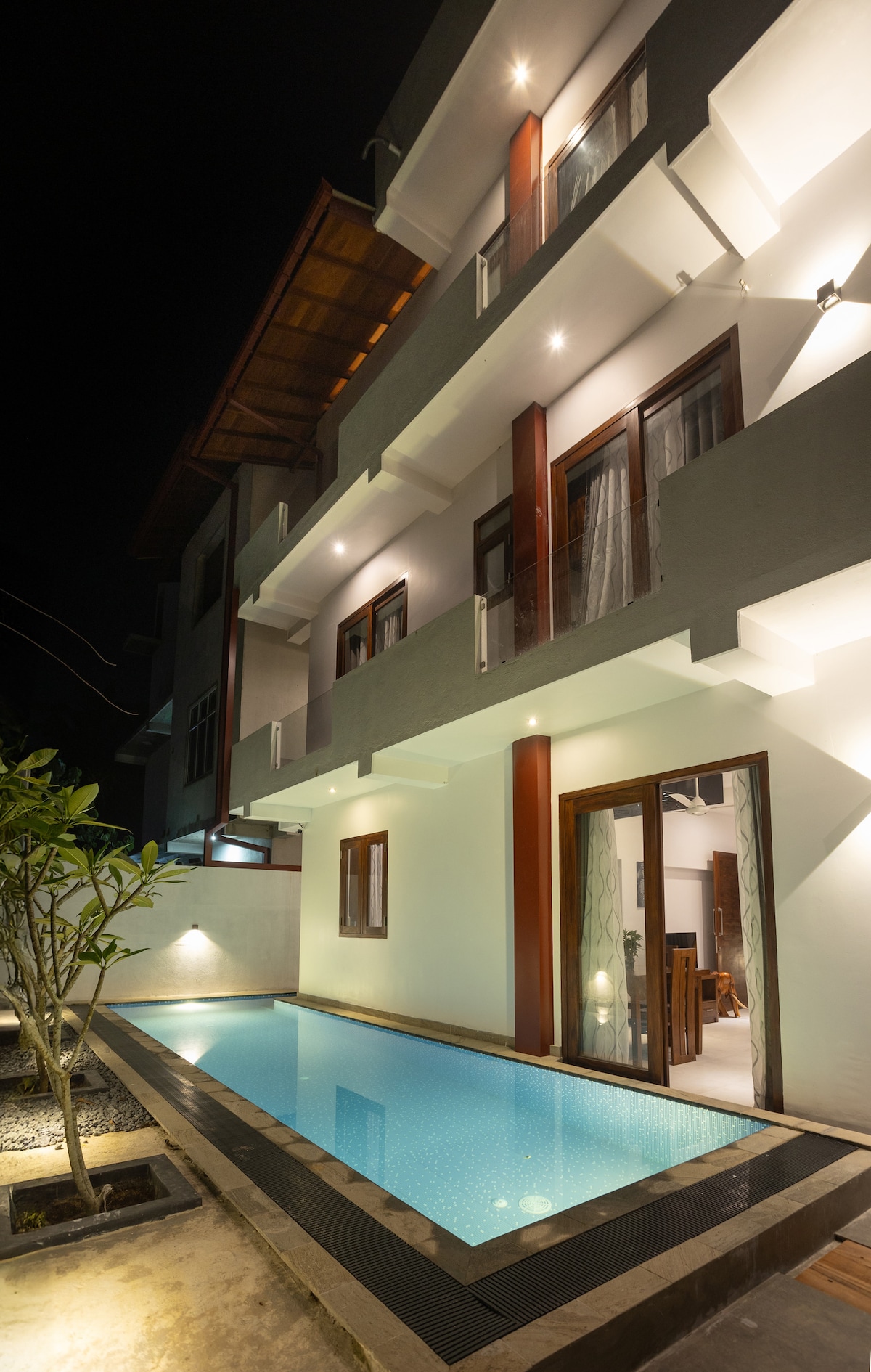 An illuminated outdoor pool is set against the backdrop of a three-story building at night. The soft glow from the lights highlights the architecture, with glass doors leading to the interior spaces. A small landscaped area features a young tree near the pool.