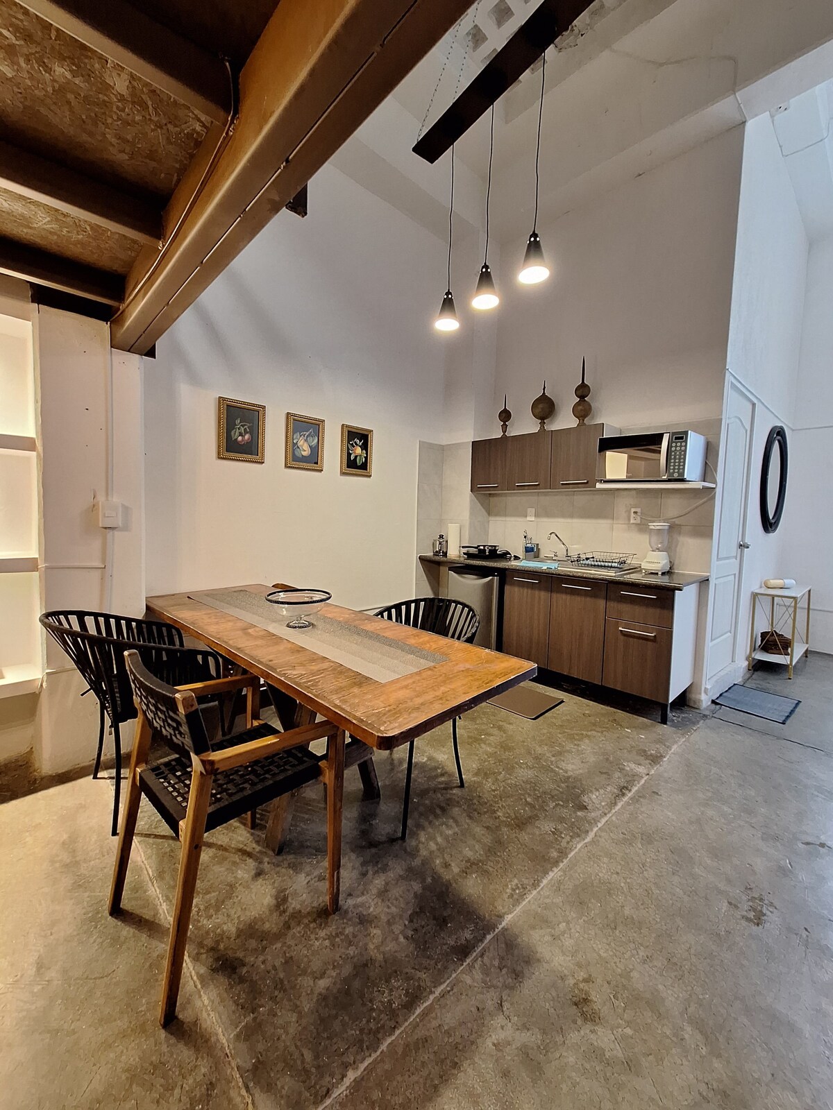 A modern kitchen area is highlighted by wooden cabinetry and stainless steel appliances. A dining table with four chairs sits in the foreground, complemented by framed artwork on the walls. Bright lighting fixtures hang above, contributing to the open and inviting space.