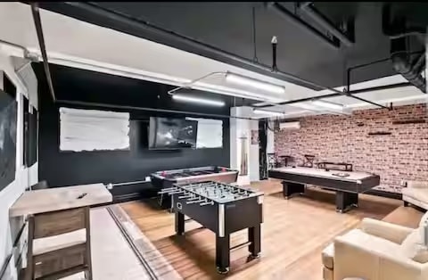 A game room is equipped with a foosball table and a billiards table, situated against a backdrop of exposed brick walls. A large screen television is mounted on the wall, with soft lighting provided by overhead fixtures. Comfortable seating is available in a neutral tone.