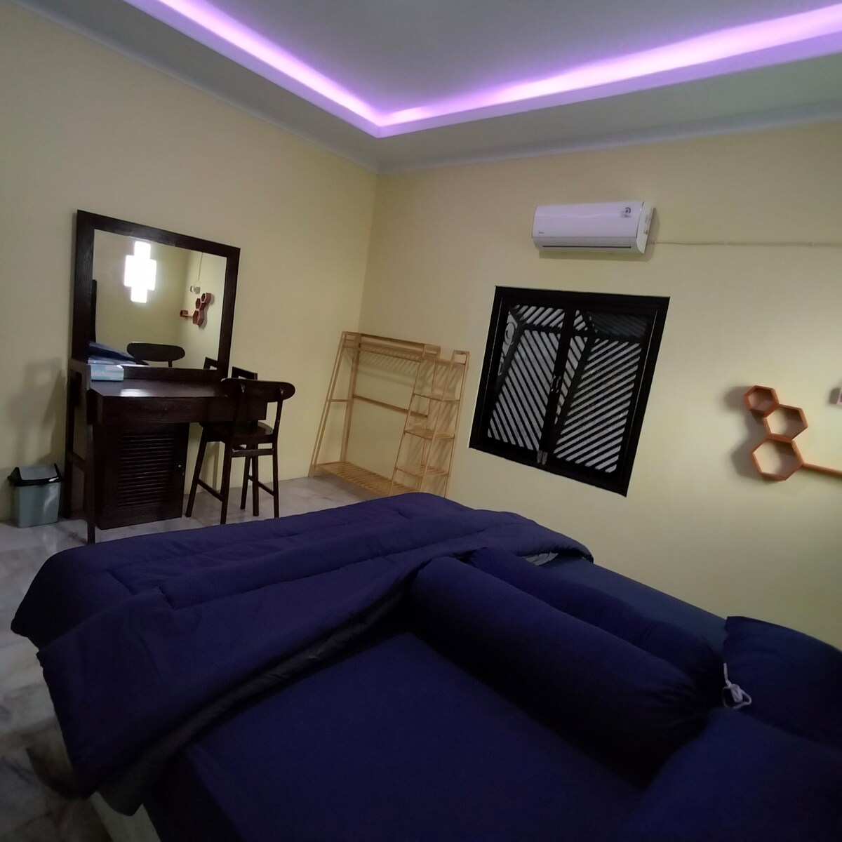 A cozy sleeping area is featured, with a spacious bed dressed in dark bedding. A wooden vanity with bar stools provides a functional workspace. Cool air conditioning is present on the wall, and a geometric wall decoration adds a modern touch. Natural light enters through a window covered by black slats.