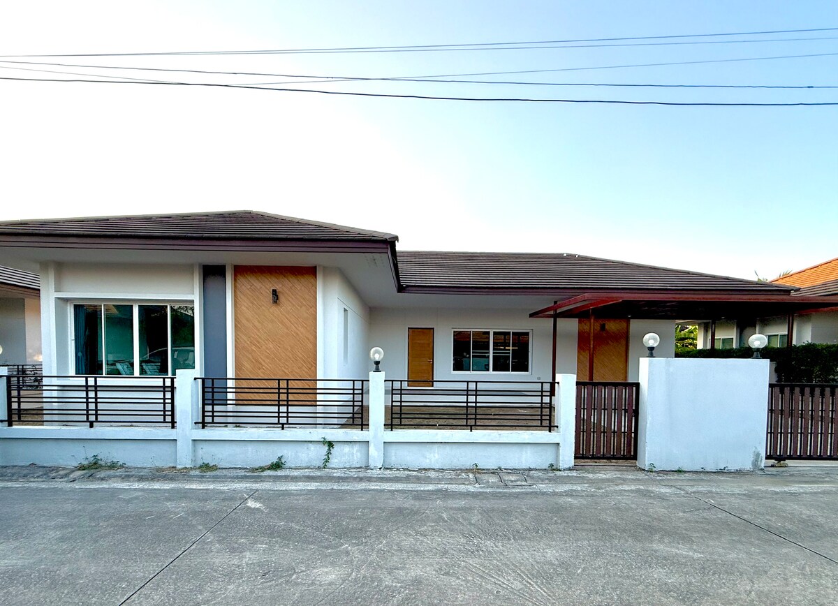 The exterior of a single-story home is presented, featuring a combination of wooden and white walls. A covered porch with simple light fixtures is visible, alongside a gated entrance. The driveway is accessible, with a well-kept garden area bordering the property.