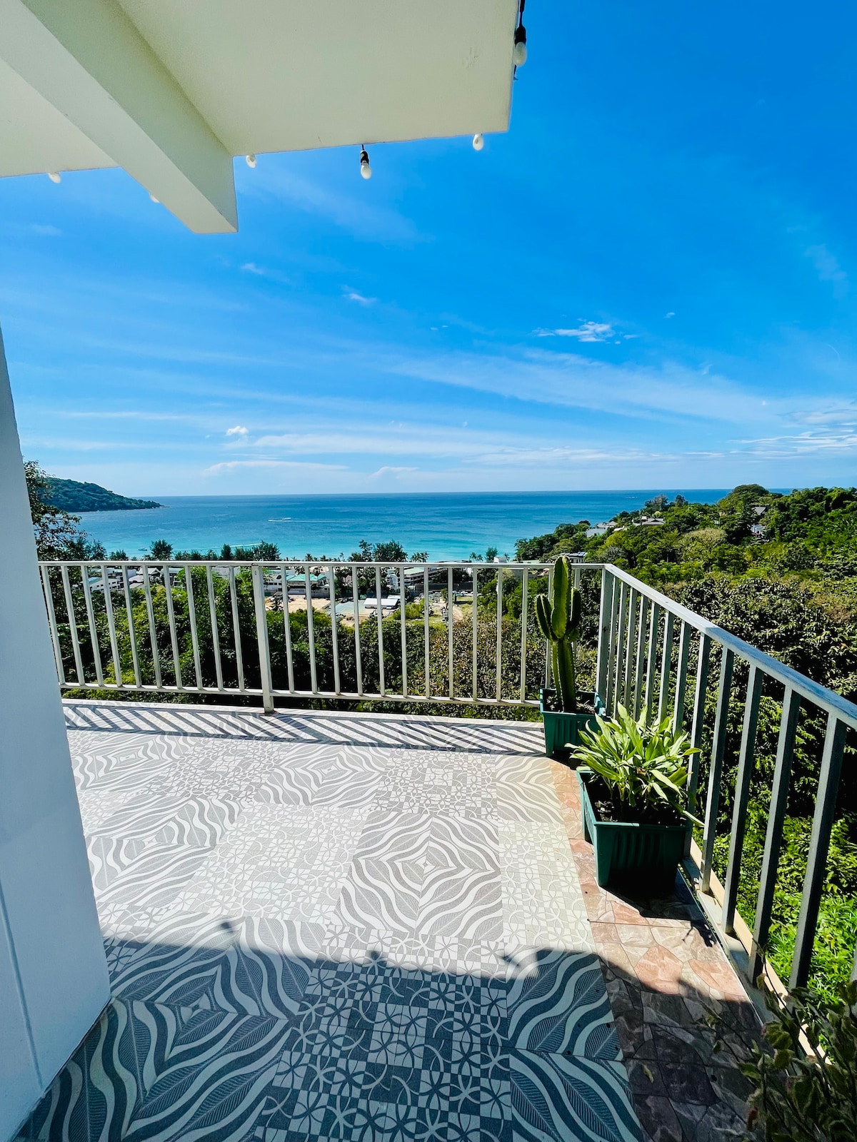 A spacious terrace is displayed, featuring patterned tiles and a railing that overlooks a scenic ocean view. Lush greenery is visible in potted plants, while the clear blue sky enhances the overall brightness of the setting.