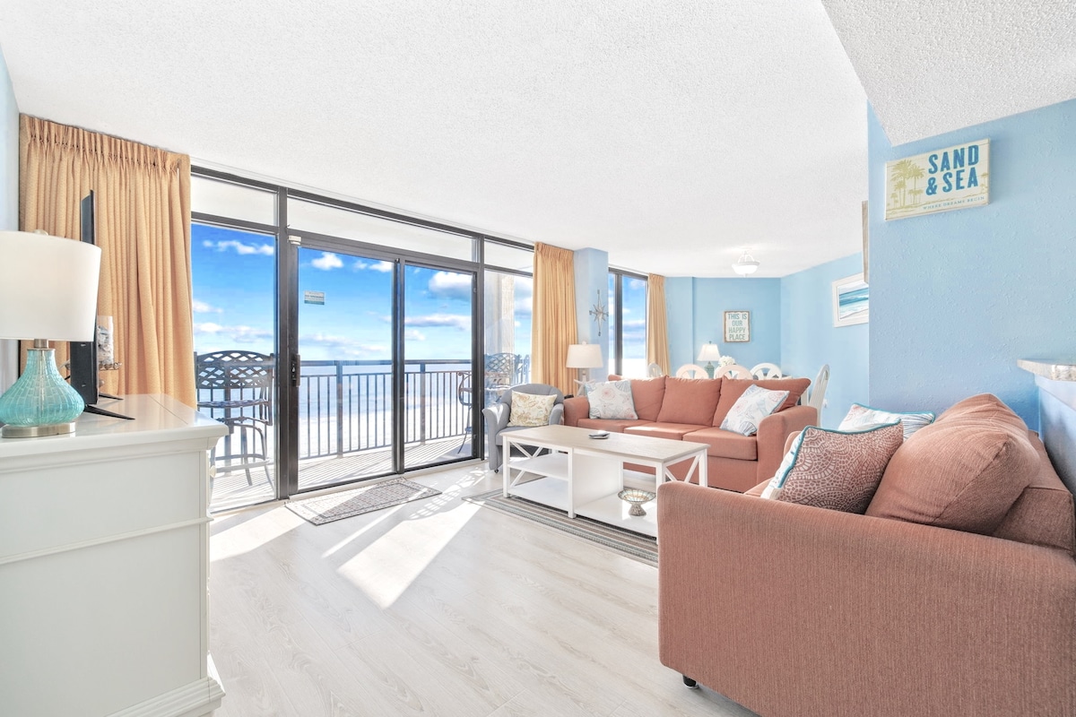 A bright and airy living area features large sliding glass doors that open to a spacious oceanfront balcony. Cozy seating is arranged around a central coffee table, with soft hues of blue and coral complementing the coastal decor. A smart TV is positioned for entertainment.