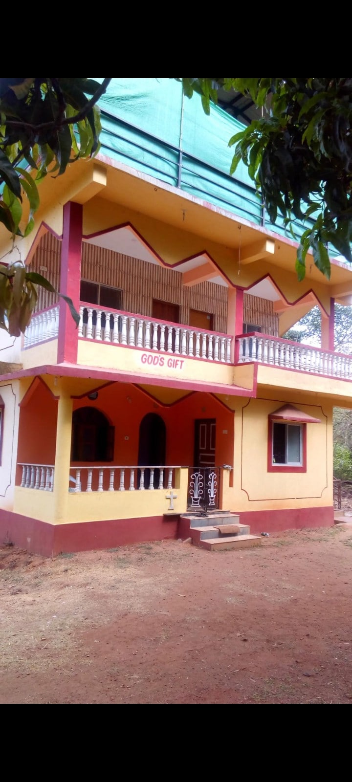 A two-story structure features a vibrant exterior with a combination of yellow and red paint. The balcony is adorned with white railings, and a set of steps leads to the main entrance. Large windows are present, contributing to a welcoming facade.