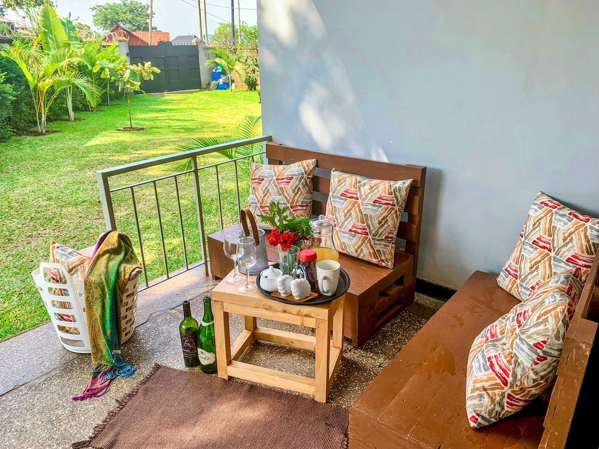 An outdoor seating area is highlighted with wooden furniture and decorative cushions. A small table is set with tea utensils and floral accents. A view of the lush green backyard is visible, enhancing the peaceful atmosphere of the space.